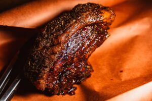 Close-up of a smoked lamb shoulder on orange butcher paper, showing the moist, dark crust and seasoned exterior.